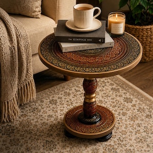 May include: A round wooden side table featuring an intricate mandala pattern in shades of brown, gold, and black. The table has a central pedestal base with decorative carvings and rests on small feet. The surface holds two books and a mug.