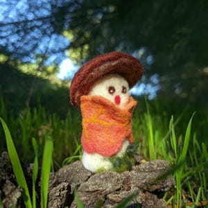 Puede incluir: Una figura de hongo de fieltro con cuerpo blanco, sombrero marrón oscuro y boca roja. La figura está envuelta en un chal con forma de hoja naranja y amarilla con costuras visibles y presenta un pequeño parche verde en la base.