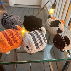 May include: Six small, round, crocheted plush cat toys in various colors and patterns. The collection includes a solid orange cat, a solid black cat, a gray tabby, a white and gray tabby, a calico, and a white cat with dark brown ears and tail.
