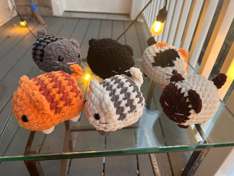 May include: Six small, round, crocheted plush cat toys in various colors and patterns. The collection includes a solid orange cat, a solid black cat, a gray tabby, a white and gray tabby, a calico, and a white cat with dark brown ears and tail.