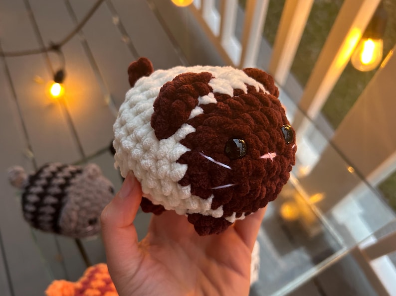 May include: A round, plush crochet guinea pig toy featuring a two-tone color pattern of dark brown and cream white yarn. The stuffed animal has two shiny black plastic eyes, a small pink stitched nose, and white yarn whiskers on its face.