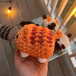 May include: A small, plush, crocheted orange cat toy with dark orange tabby stripes, a curled tail, and white paws. The cat has black bead eyes and white whiskers. In the background, four other similar crocheted plush toys are visible.