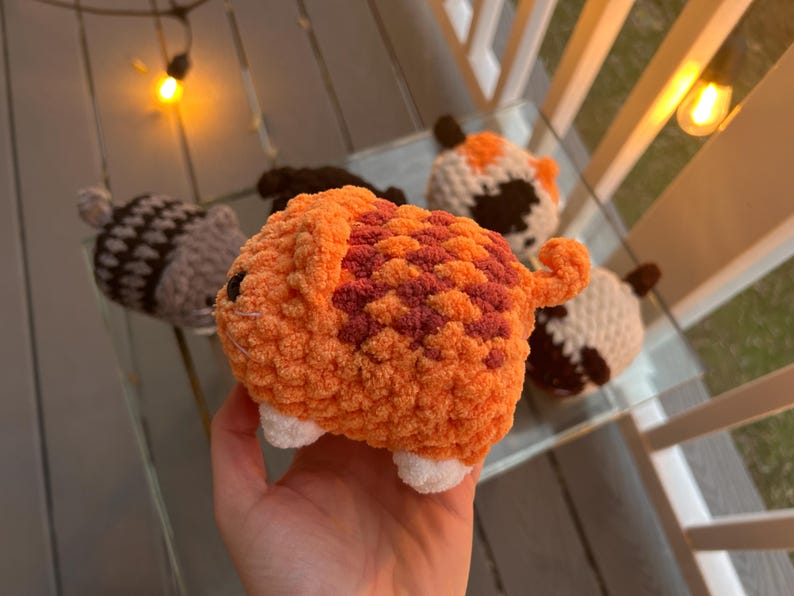 May include: A small, plush, crocheted orange cat toy with dark orange tabby stripes, a curled tail, and white paws. The cat has black bead eyes and white whiskers. In the background, four other similar crocheted plush toys are visible.