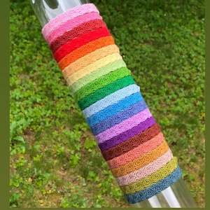 May include: A stack of colourful woven bracelets in a rainbow pattern. The bracelets are made of a textured material and have a diamond pattern.