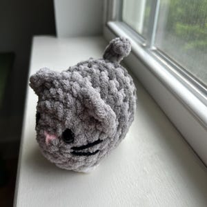 May include: A small, round, gray crocheted cat plushie made from soft, chunky yarn. The cat features two pointed ears, a small pink stitched nose, black plastic safety eyes, and black embroidered whiskers, with a tiny tail on its back.