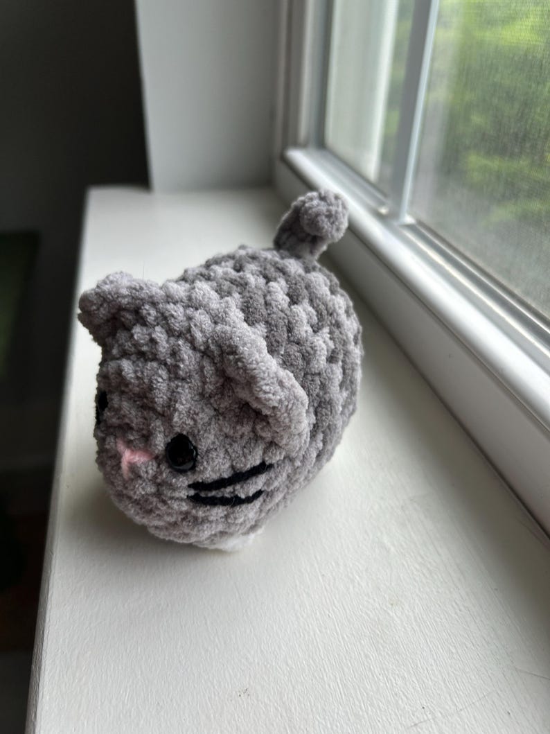 May include: A small, round, gray crocheted cat plushie made from soft, chunky yarn. The cat features two pointed ears, a small pink stitched nose, black plastic safety eyes, and black embroidered whiskers, with a tiny tail on its back.