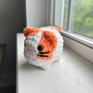 May include: A small, plush, crocheted cat figure made of soft white and orange yarn. The cat has round black plastic eyes, a small pink stitched nose, and two horizontal black yarn stripes on each cheek, with a rounded body and small ears.