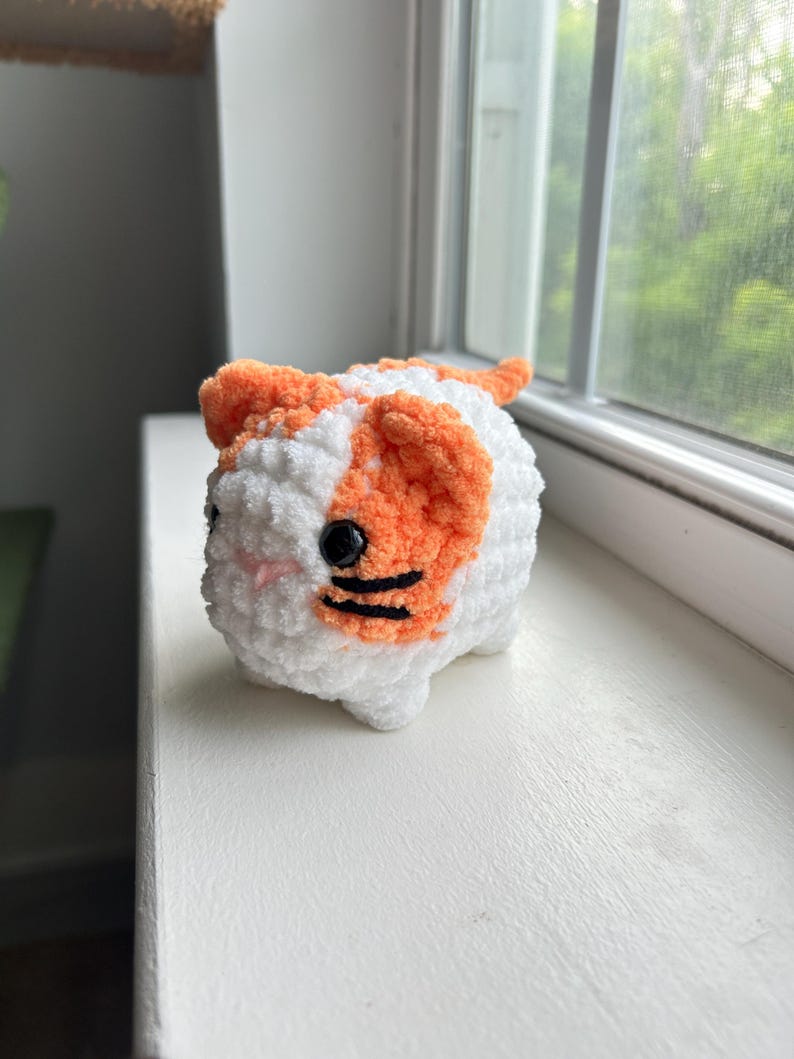 May include: A small, plush, crocheted cat figure made of soft white and orange yarn. The cat has round black plastic eyes, a small pink stitched nose, and two horizontal black yarn stripes on each cheek, with a rounded body and small ears.