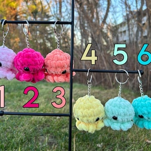 May include: Six crocheted octopus keychains in various colors, including pink, coral, yellow, light blue, and teal. The keychains are hanging on a black metal stand with numbers 1 through 6 labeled below each keychain.