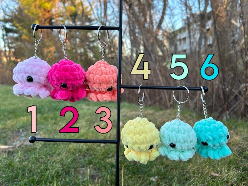 May include: Six crocheted octopus keychains in various colors, including pink, coral, yellow, light blue, and teal. The keychains are hanging on a black metal stand with numbers 1 through 6 labeled below each keychain.