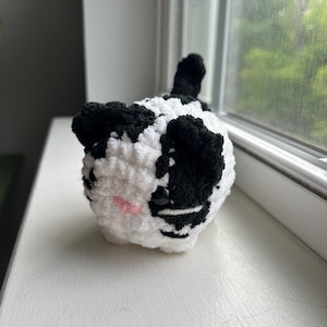 May include: A small, round, crocheted plush cat with a white and black spotted pattern. The cat features black ears, a small pink stitched nose, and two dark, glossy plastic eyes. A short, upright black tail is visible on the back of the figure.