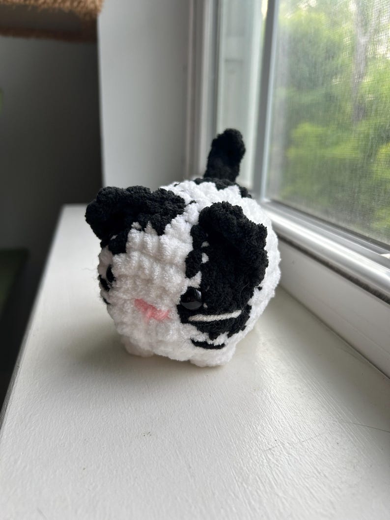 May include: A small, round, crocheted plush cat with a white and black spotted pattern. The cat features black ears, a small pink stitched nose, and two dark, glossy plastic eyes. A short, upright black tail is visible on the back of the figure.