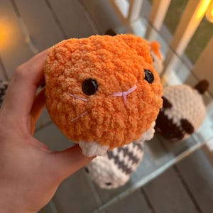 May include: A small, round, orange crocheted plush toy with a textured, fuzzy yarn surface. It features two shiny black plastic eyes, a small pink stitched nose, and white yarn accents at the base. The plush has a soft, spherical shape.