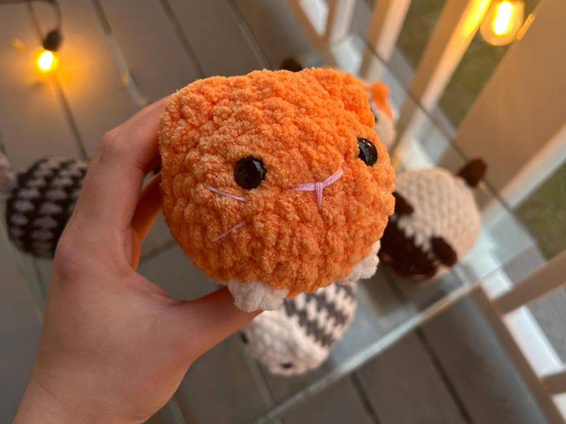 May include: A small, round, orange crocheted plush toy with a textured, fuzzy yarn surface. It features two shiny black plastic eyes, a small pink stitched nose, and white yarn accents at the base. The plush has a soft, spherical shape.