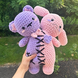 May include: A handmade crochet plush toy featuring a bear and a bunny, joined together. The bear is lavender, the bunny is pink. Both have black stitched details and a heart-shaped bow. The toy is held in front of a green background.