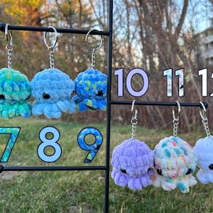 May include: Twelve crocheted octopus keychains in various colors, including blue, green, purple, and white. The keychains are displayed on a metal rack with numbers 7 through 12.