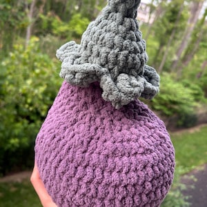 May include: A plush, hand-crocheted eggplant toy made from soft, textured purple yarn for the main body and a thick, dark green yarn for the stem and leafy top. The vegetable-shaped item features a rounded base and a pointed, textured stem.
