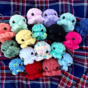 May include: A collection of crocheted octopus keychains in various colors, including pink, purple, blue, green, yellow, white, red, and black. The octopus keychains are arranged on a plaid fabric background.