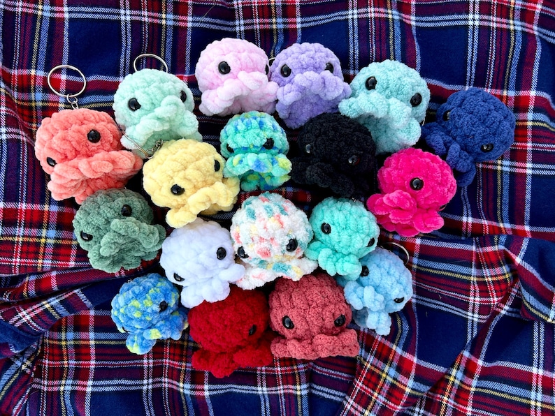 May include: A collection of crocheted octopus keychains in various colors, including pink, purple, blue, green, yellow, white, red, and black. The octopus keychains are arranged on a plaid fabric background.