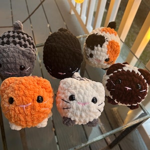 May include: Six small, round, crocheted plush cat toys in various colors and patterns. The collection includes a gray checkered cat, a solid black cat, a calico cat, an orange cat, a white cat with gray paws, and a brown and white cat.