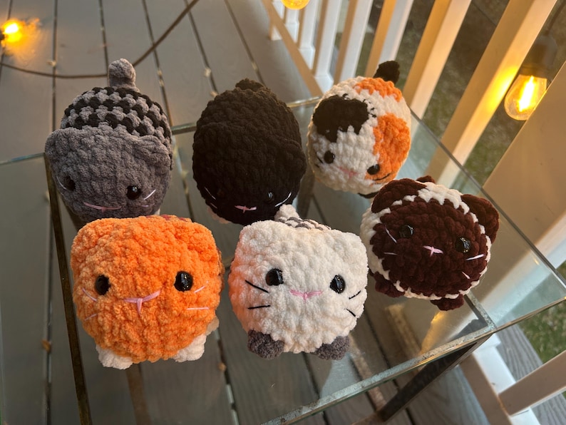 May include: Six small, round, crocheted plush cat toys in various colors and patterns. The collection includes a gray checkered cat, a solid black cat, a calico cat, an orange cat, a white cat with gray paws, and a brown and white cat.