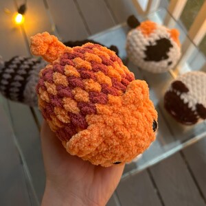 May include: A plush, hand-crocheted toy shaped like an acorn with a textured, orange and maroon cap and a solid orange body. The toy features a small stem on top and dark, round eyes. Other similar crocheted acorn toys are visible in the background.