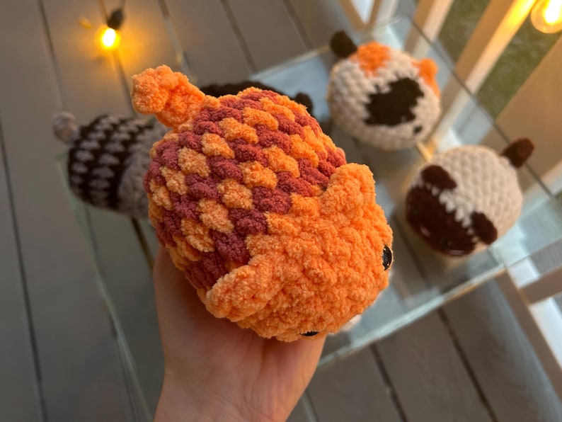 May include: A plush, hand-crocheted toy shaped like an acorn with a textured, orange and maroon cap and a solid orange body. The toy features a small stem on top and dark, round eyes. Other similar crocheted acorn toys are visible in the background.