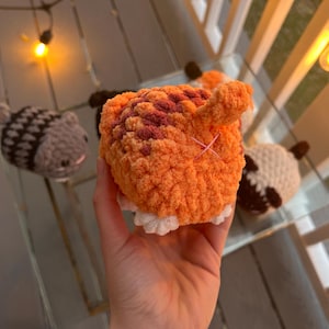 May include: A small, round, orange and brown crocheted plush toy shaped like an animal rear, featuring a small upright tail and a pink stitched cross detail. The plush has white crocheted feet at the base and a textured, soft yarn construction.