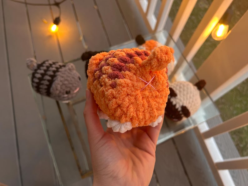 May include: A small, round, orange and brown crocheted plush toy shaped like an animal rear, featuring a small upright tail and a pink stitched cross detail. The plush has white crocheted feet at the base and a textured, soft yarn construction.