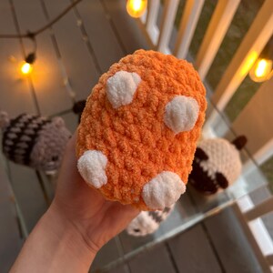 May include: A small, round, orange plush toy made of soft, fuzzy yarn. The object features four distinct, white, rounded, textured patches on its surface, resembling small paws or pads. The material has a thick, looped, and tactile knit texture.