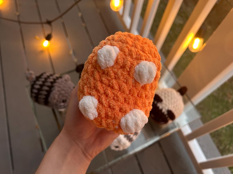 May include: A small, round, orange plush toy made of soft, fuzzy yarn. The object features four distinct, white, rounded, textured patches on its surface, resembling small paws or pads. The material has a thick, looped, and tactile knit texture.