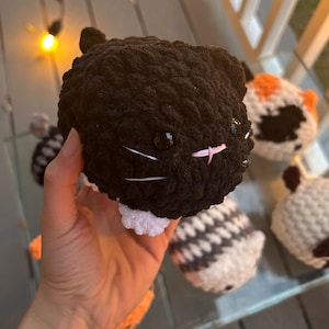 May include: A round, black crocheted cat plush toy with white paws, small black ears, shiny black plastic eyes, and a stitched pink nose with white whiskers. The plush has a textured, yarn-based surface and is held in a hand, surrounded by other similar crocheted animal plush toys.