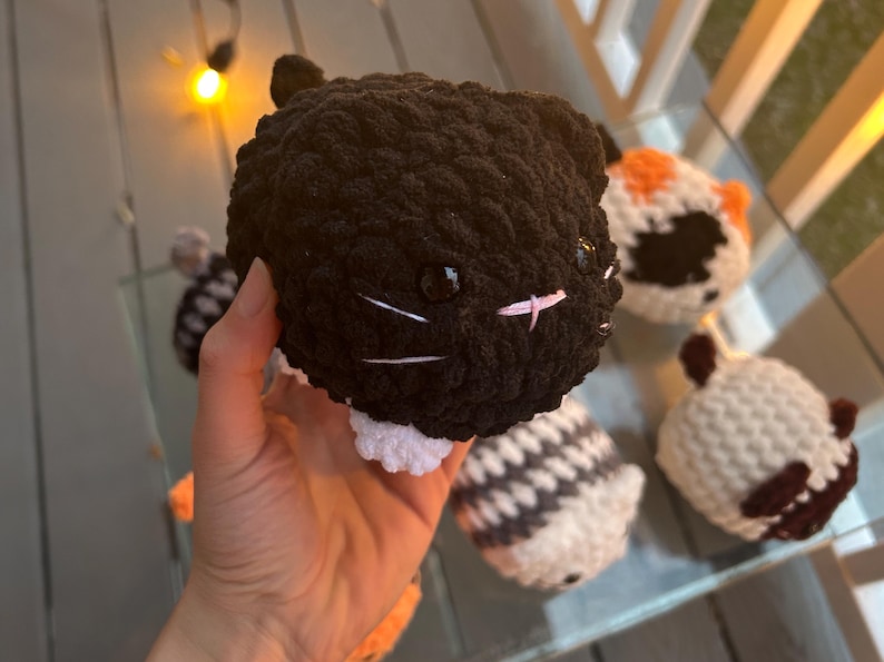 May include: A round, black crocheted cat plush toy with white paws, small black ears, shiny black plastic eyes, and a stitched pink nose with white whiskers. The plush has a textured, yarn-based surface and is held in a hand, surrounded by other similar crocheted animal plush toys.