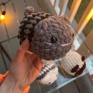 May include: A small, round, crocheted plush toy shaped like a cat with dark gray and black yarn, white paws, black plastic eyes, and embroidered white whiskers and a pink nose. The plush has a textured, soft appearance with a small tail.