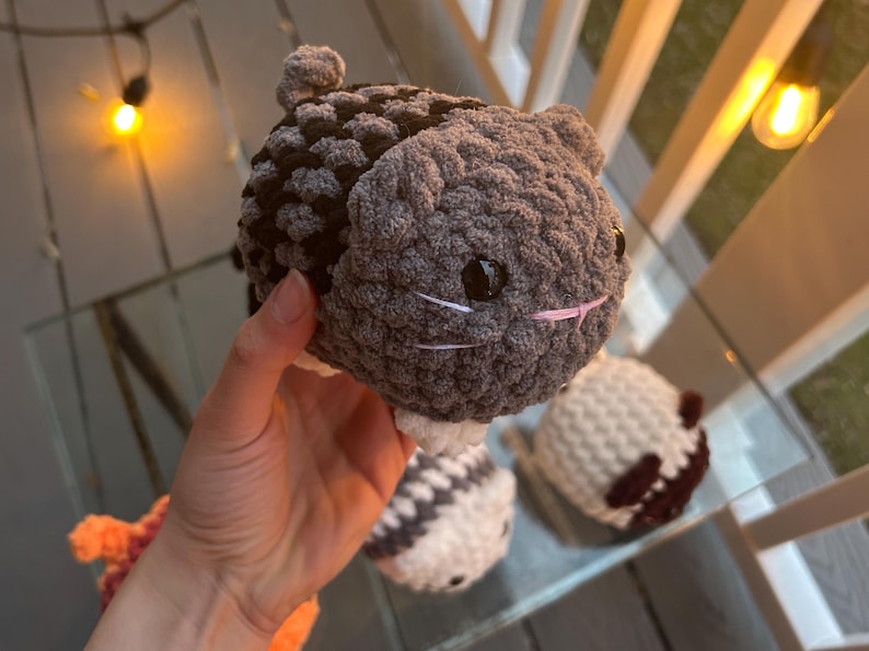 May include: A small, round, crocheted plush toy shaped like a cat with dark gray and black yarn, white paws, black plastic eyes, and embroidered white whiskers and a pink nose. The plush has a textured, soft appearance with a small tail.