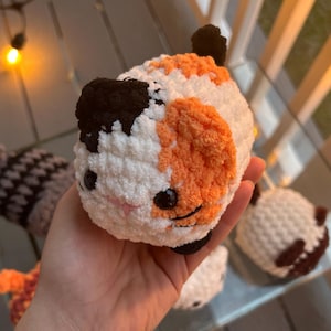 May include: A small, round, handmade crochet plush toy shaped like a calico cat. The plush features soft white, orange, and black yarn, with two black plastic safety eyes, a small pink stitched nose, and black stitched whiskers on its face.