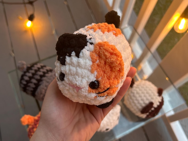 May include: A small, round, handmade crochet plush toy shaped like a calico cat. The plush features soft white, orange, and black yarn, with two black plastic safety eyes, a small pink stitched nose, and black stitched whiskers on its face.