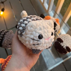 May include: A round, plush, white and gray crocheted cat toy with black plastic eyes and embroidered whiskers. The cat has a small white tail and gray paws. Several other similar crocheted animal toys in various colors are visible in the background.