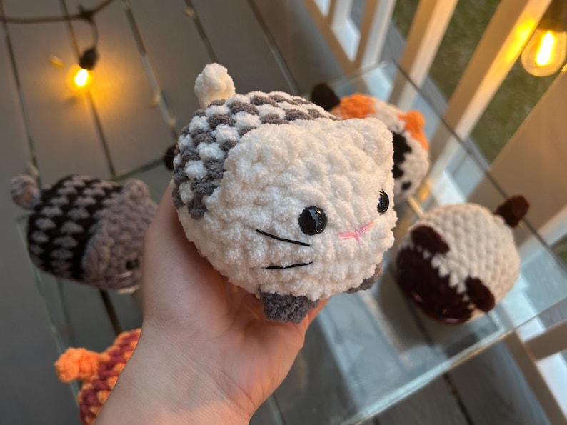 May include: A round, plush, white and gray crocheted cat toy with black plastic eyes and embroidered whiskers. The cat has a small white tail and gray paws. Several other similar crocheted animal toys in various colors are visible in the background.