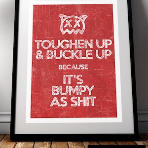 May include: Framed art print with a red background and white text that reads "TOUGHEN UP & BUCKLE UP BECAUSE IT'S BUMPY AS SHIT." The print features a distressed texture and a cartoon face at the top. The frame is black.