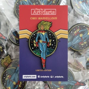 Op de afbeelding: Een gelimiteerde editie emaille pin met een superheld in een blauw en rood kostuum met een gele ster op de borst. De pin is geplaatst tegen een zwarte achtergrond met sterren en een planeet. De pin is gelabeld "CM01 MARVELLOUS" en "PINS BY ARTYFARTS".