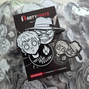 May include: A black and white enamel pin set featuring two characters, one with a cross earring and one wearing a wide-brimmed hat and round glasses. The design includes the text "HELLO DEAR FELLOW" and a flame icon on a black display card.