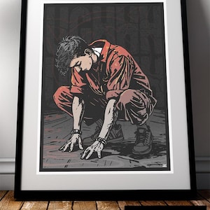 May include: Framed art print depicting a person in a red jumpsuit, crouching with hands outstretched. The artwork has a dark, textured background with graffiti-style elements. The frame is black, and the print is displayed on a wooden surface.
