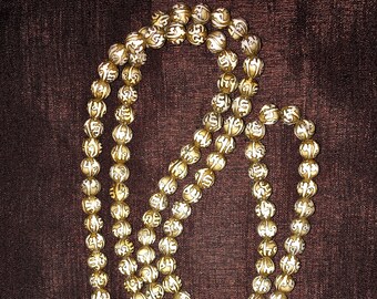 May include: A white mala bead necklace with gold lettering on each bead. The necklace is on a brown fabric background and has a red tassel.