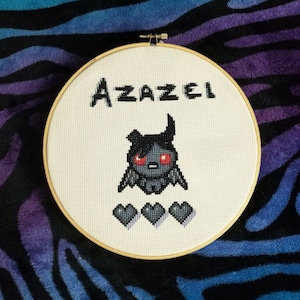 Op de afbeelding: Een witte borduurring met een kruissteekontwerp van een zwart en rood cartoonfiguur met de naam "Azazel" erboven en drie zwarte harten eronder.