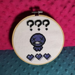 May include: A purple character with a question mark above its head and three hearts below it. The character is embroidered on a white fabric with a wooden hoop.