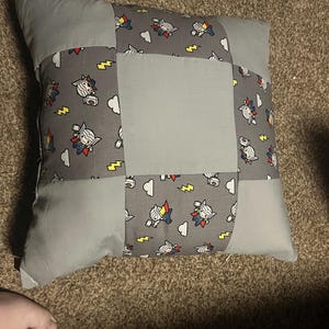 May include: A square gray pillow with a patchwork design. The pillow features a central gray square surrounded by gray fabric panels. The panels are decorated with a repeating pattern of a cartoon character, clouds, and lightning bolts.