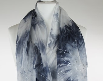 Op de afbeelding: Een witte zijden sjaal met een blauw en wit tie-dye patroon en zilveren bloemenborduurwerk.