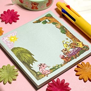 May include: A square light blue sticky note pad featuring a printed border of Winnie the Pooh characters including Piglet, Tigger, and Pooh Bear having a picnic under a tree, with a yellow sun in the corner and a yellow multi-color ink pen nearby.
