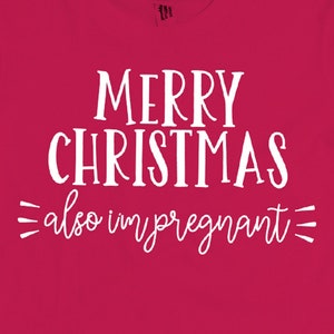 以下が含まれることがあります： 白い文字で「メリークリスマス、妊娠中」と書かれた赤いTシャツ。Tシャツには「M」の文字が書かれた白いタグが付いています。Tシャツは木製の背景の上に置かれています。画像の右下には黒い帽子に「mad hatter MOMMY」の文字が書かれています。